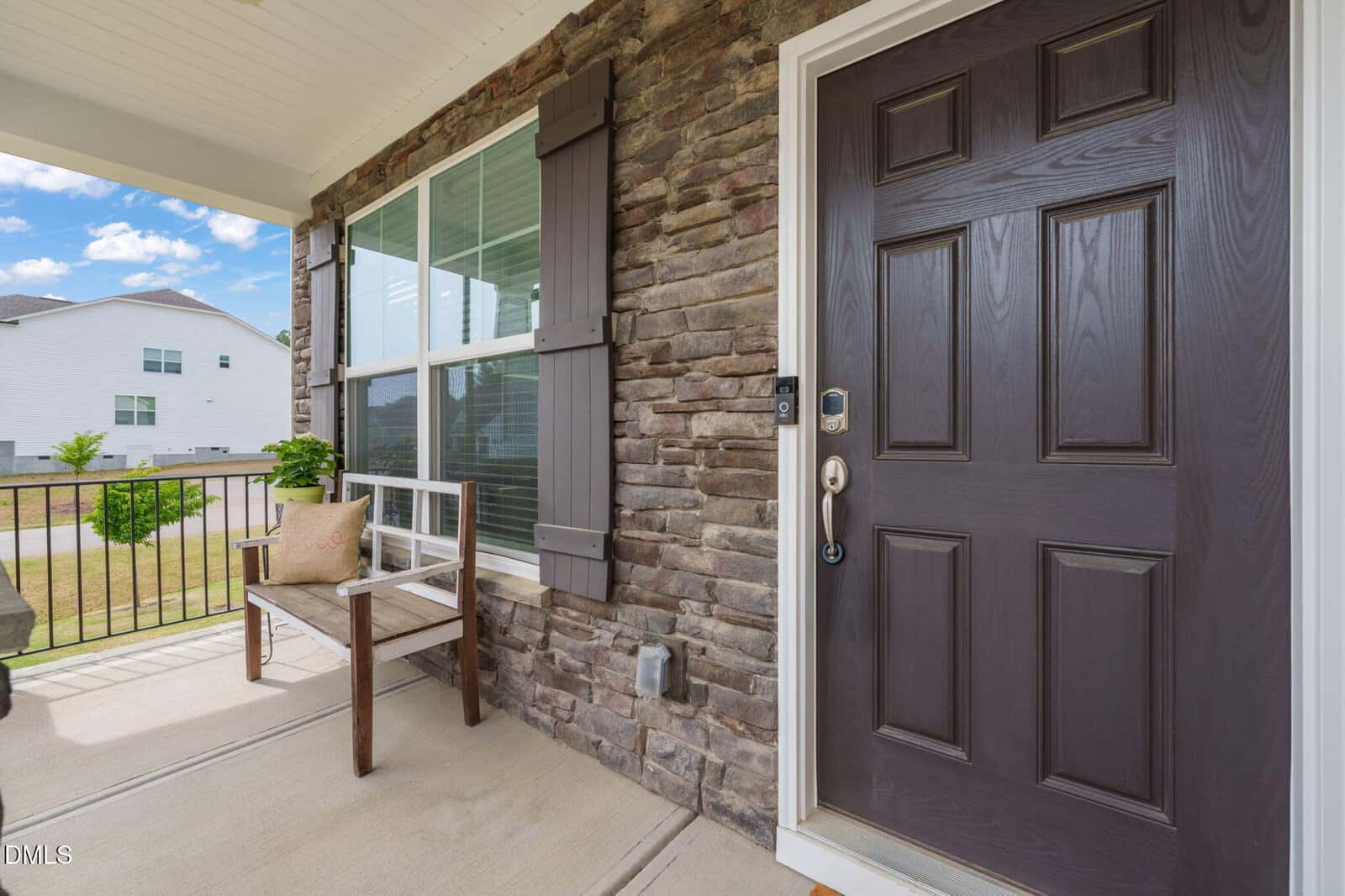 A front porch with a dark brown door, stone wall, large window with shutters, a wooden bench with a cushion, and a potted plant. A neighboring house and blue sky with clouds are visible in the background.