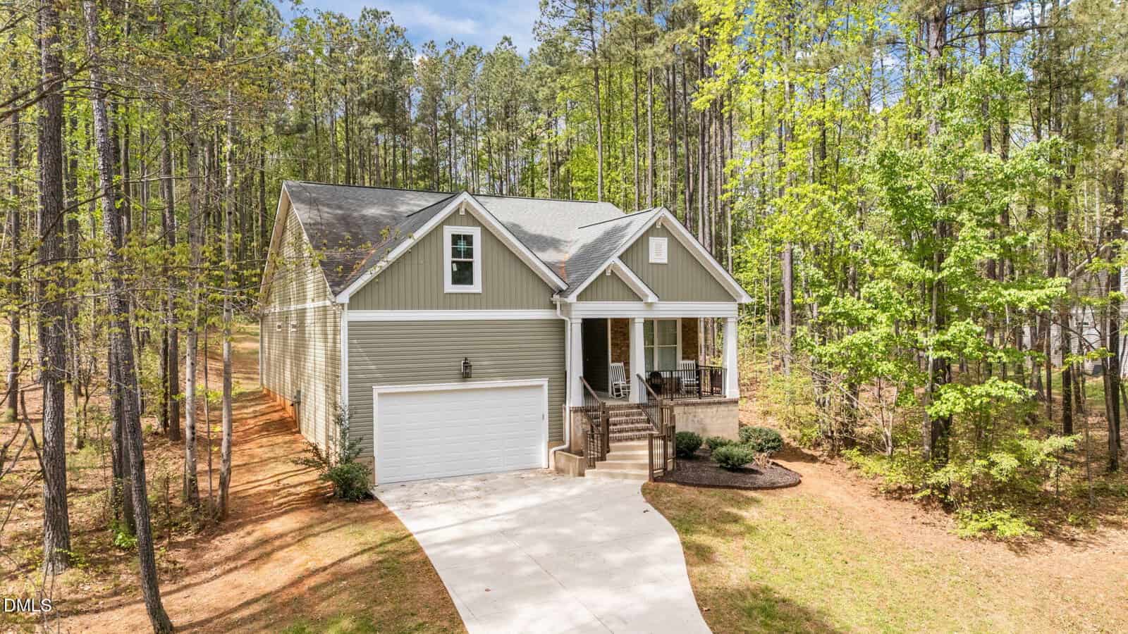 A two-story house with green siding and a front porch sits among tall trees. The home has a white garage door, concrete driveway, and minimal landscaping. Forest surrounds the property on three sides.
