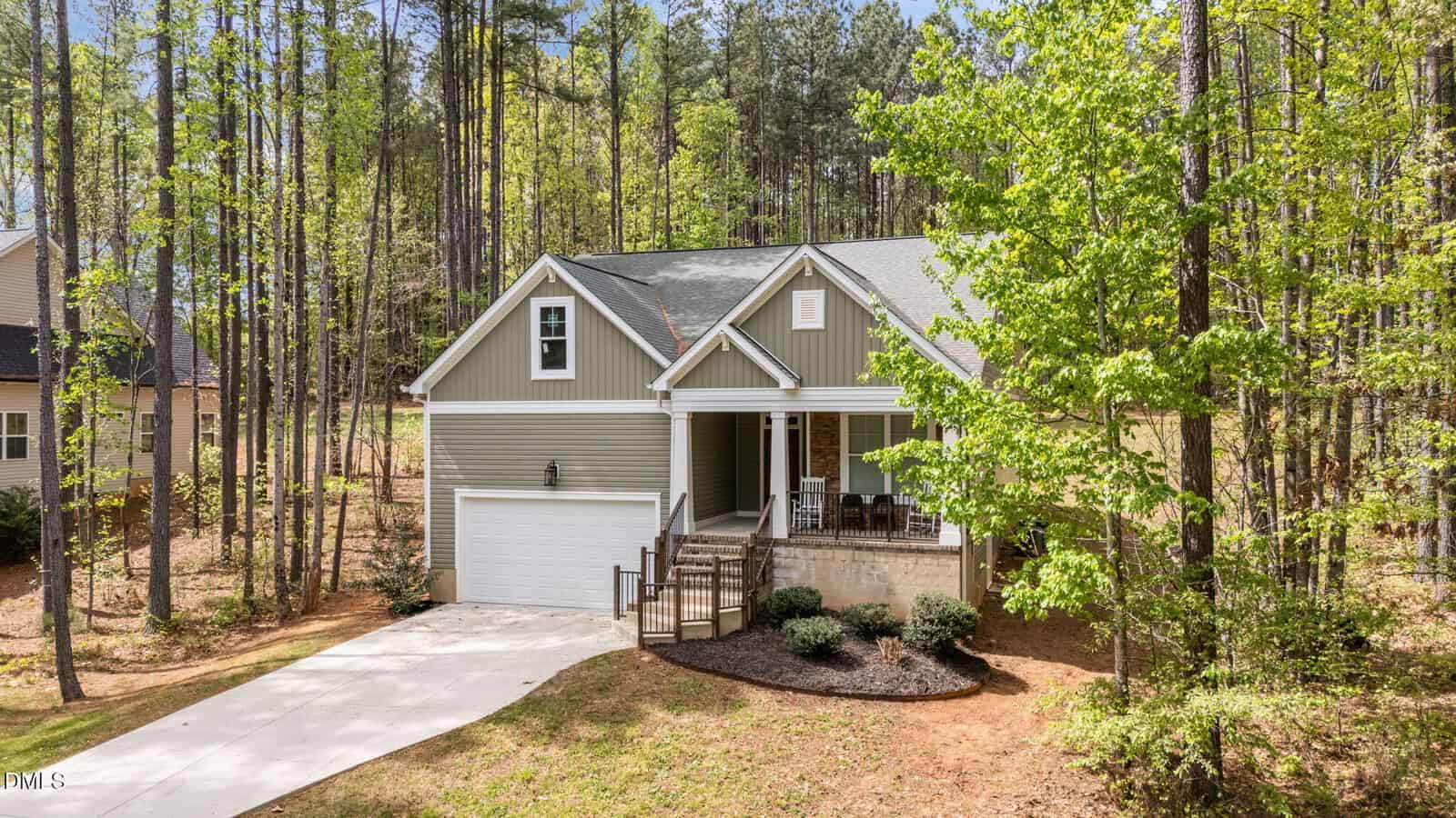 A two-story house with green siding, a front porch, and an attached garage sits among tall trees. A concrete driveway leads to the garage, and landscaped shrubs are in front of the porch.