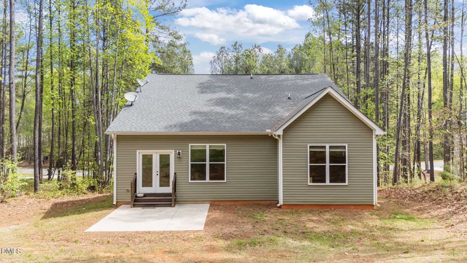 Single-story house with green siding, a gray shingled roof, two front windows, a glass double-door entrance, small front steps, and a concrete path, surrounded by tall trees and a natural landscape.