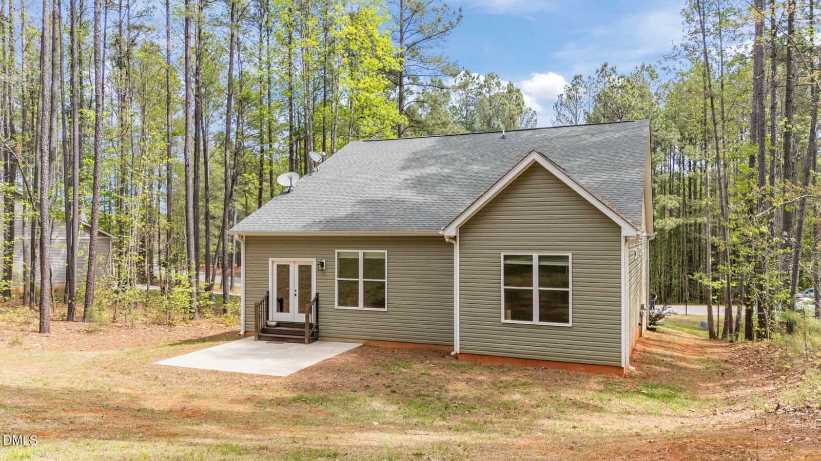 A small, single-story house with beige siding and a gray roof is set on a grassy lot surrounded by tall pine trees. There is a small porch and two large windows on the front.