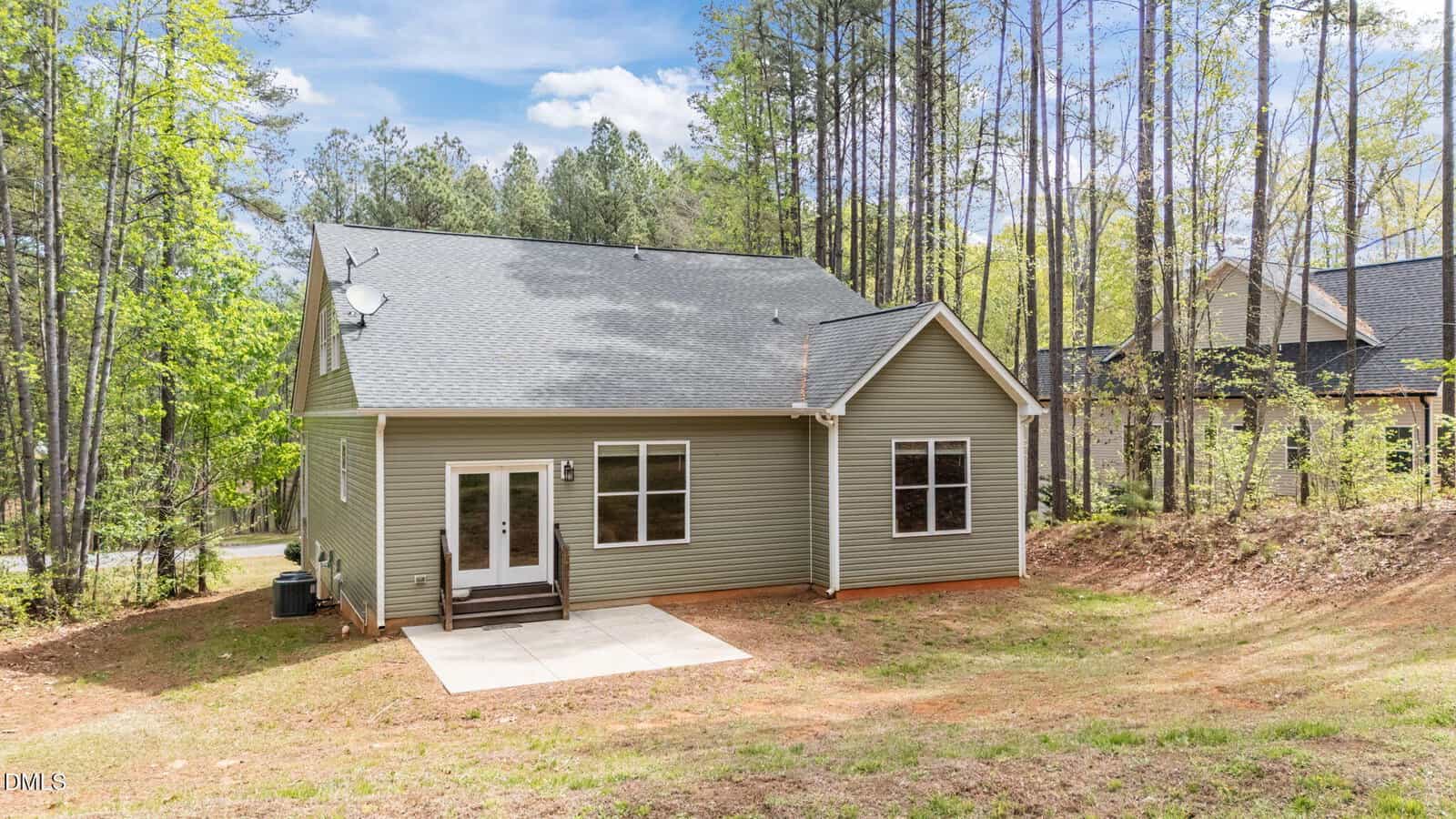 A light green house with a gray roof sits in a wooded area. There are double doors opening to a small concrete patio, large windows, and trees surrounding the home under a blue sky with clouds.