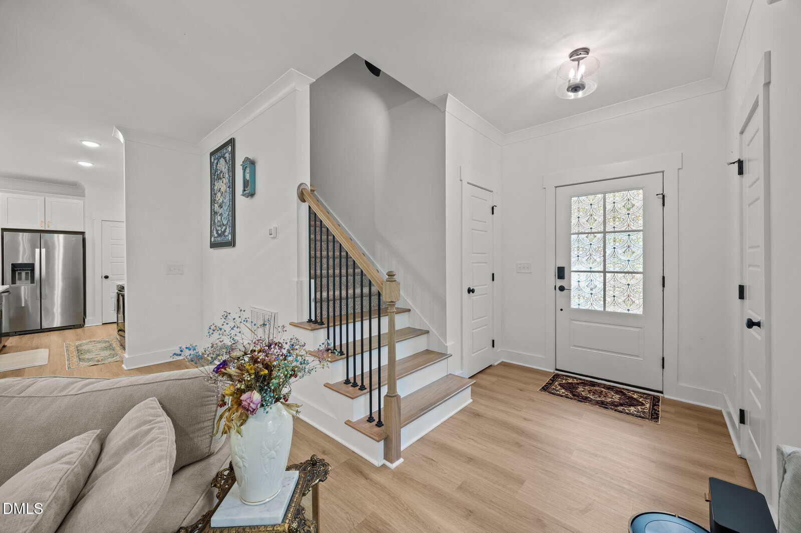 A bright entryway with light wood floors, a white front door with patterned glass, a staircase with wooden handrail and black balusters, and a vase of flowers on a side table next to a beige couch.