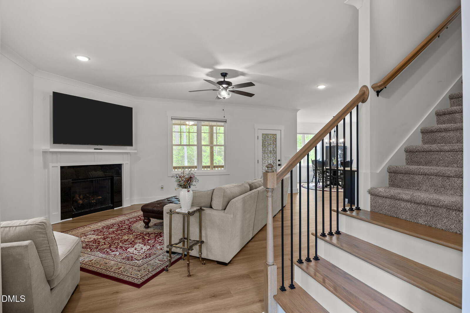 A bright living room with a beige sofa, a fireplace with a TV above it, a red patterned rug, and a staircase with wooden steps and black railings leading upstairs. Large windows let in natural light.