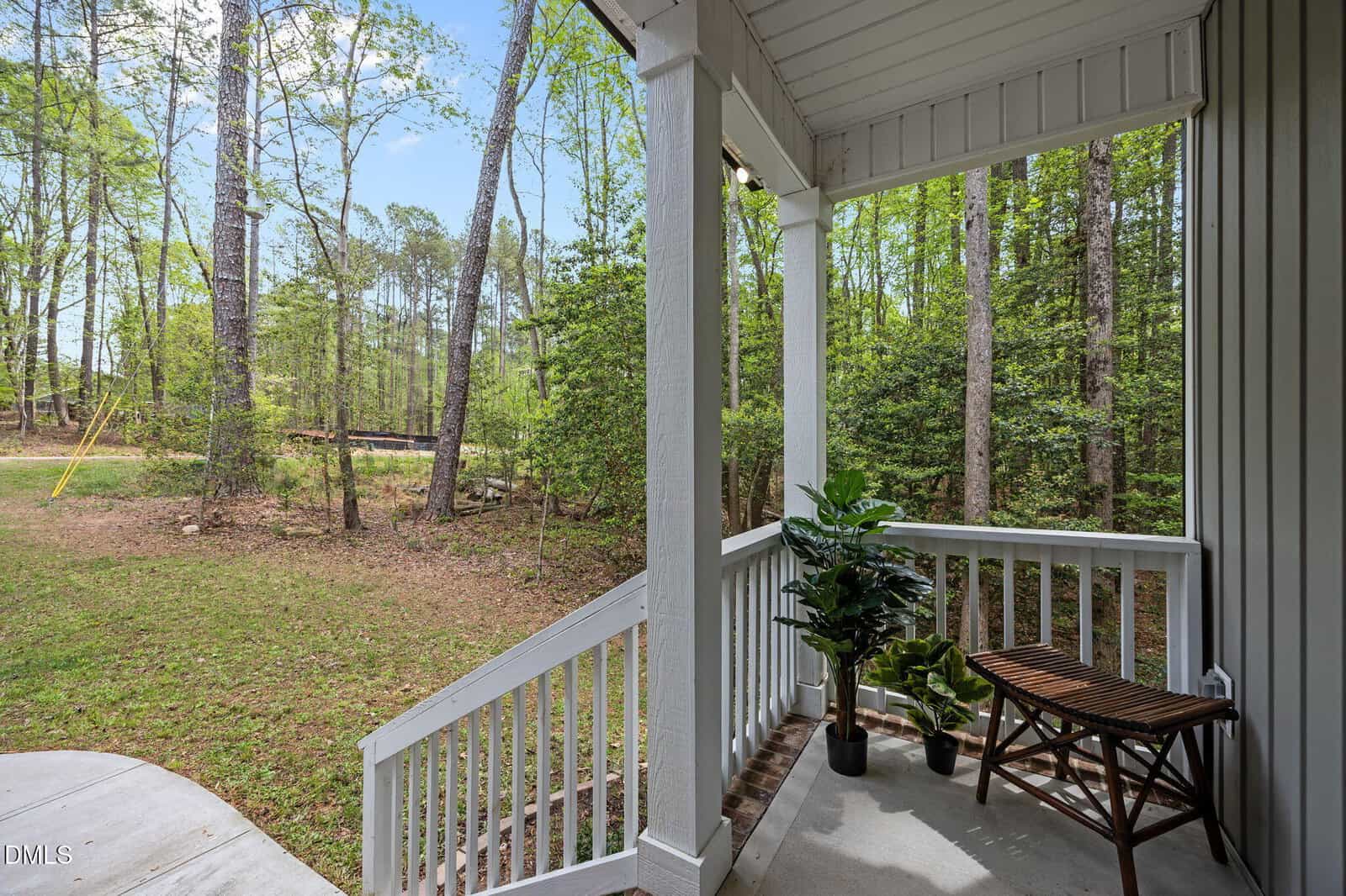 A small covered porch with white railings, a brown wooden bench, and two potted green plants overlooks a grassy yard bordered by tall trees and a forest. Steps lead down from the porch to the ground.