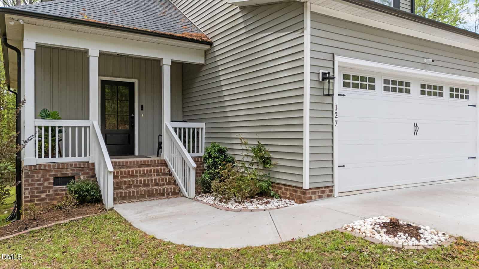 A modern gray house with white trim, a front porch with brick steps, a black door, white railings, and a two-car garage with decorative handles. Small landscaped areas with white stones and green shrubs line the walkway.