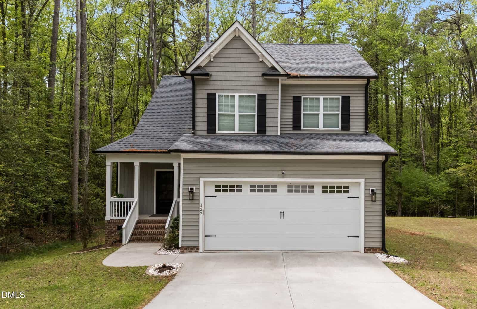 A two-story gray house with white trim and black shutters, featuring a front porch, double garage, and surrounded by trees and greenery. A concrete driveway leads up to the house.