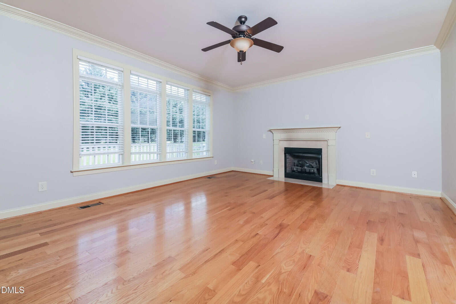 A spacious, empty living room with light hardwood floors, pale blue walls, large windows with white shutters, a ceiling fan, and a white fireplace with a mantel.