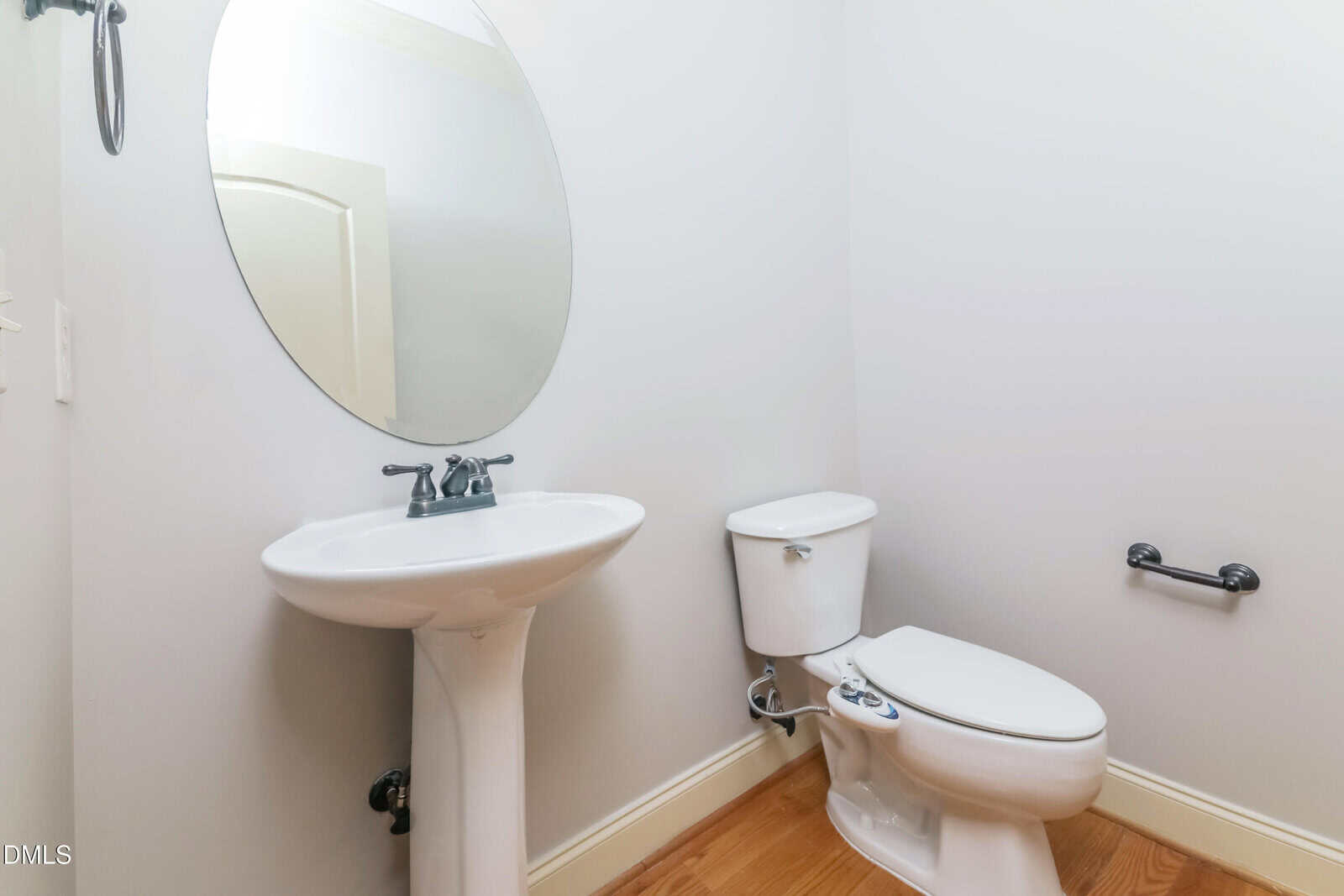A small bathroom with a pedestal sink, an oval mirror above it, a toilet, a grab bar on the wall, and light-colored walls with a wood floor.