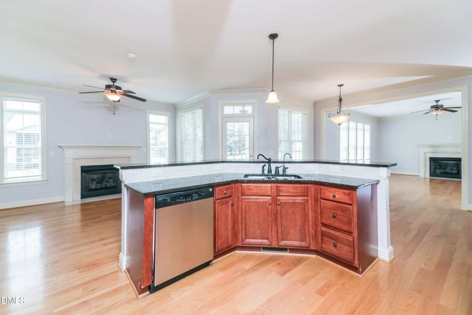 Open kitchen with dark wood cabinets, a sink, and stainless steel dishwasher, facing a spacious living area with hardwood floors, abundant windows, and two ceiling fans above separate fireplaces.