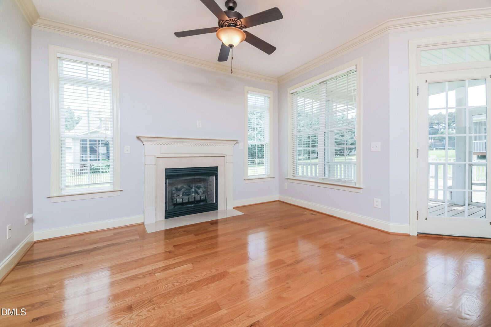 Bright living room with hardwood floors, a white fireplace, large windows with blinds, light purple walls, and a ceiling fan with lights. A door leads to a sunroom or porch area.