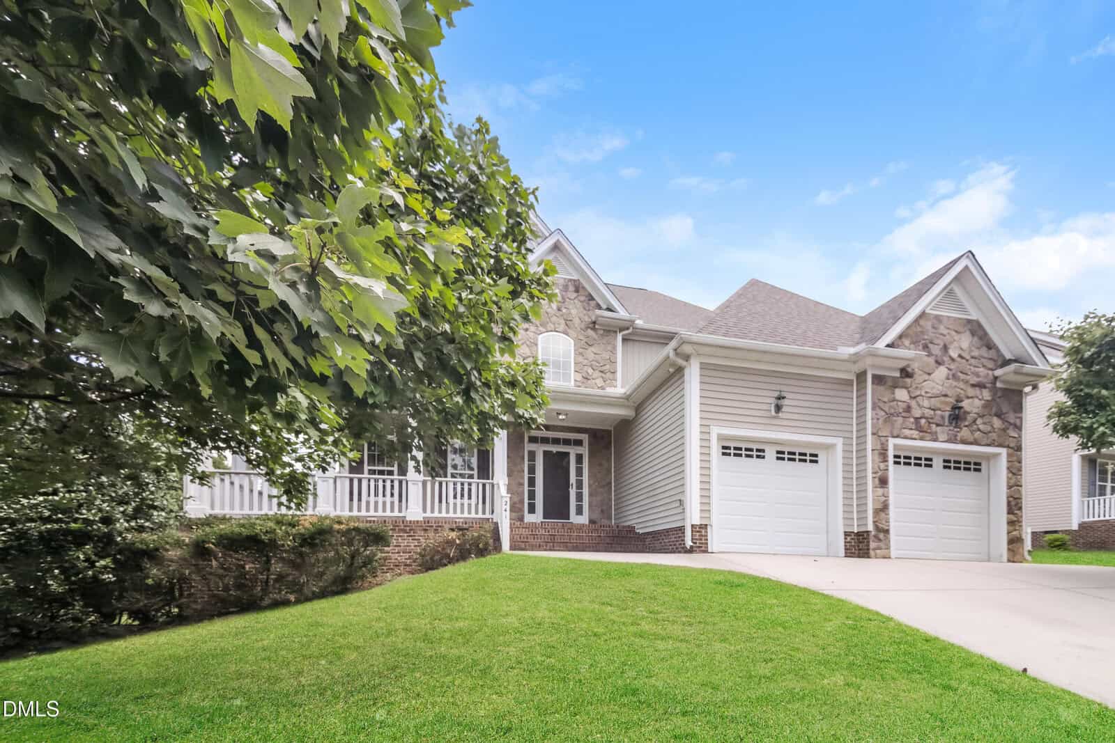 Two-story suburban house with stone and siding exterior, two white garage doors, a covered front porch, and well-maintained green lawn, set against a bright blue sky with scattered clouds.