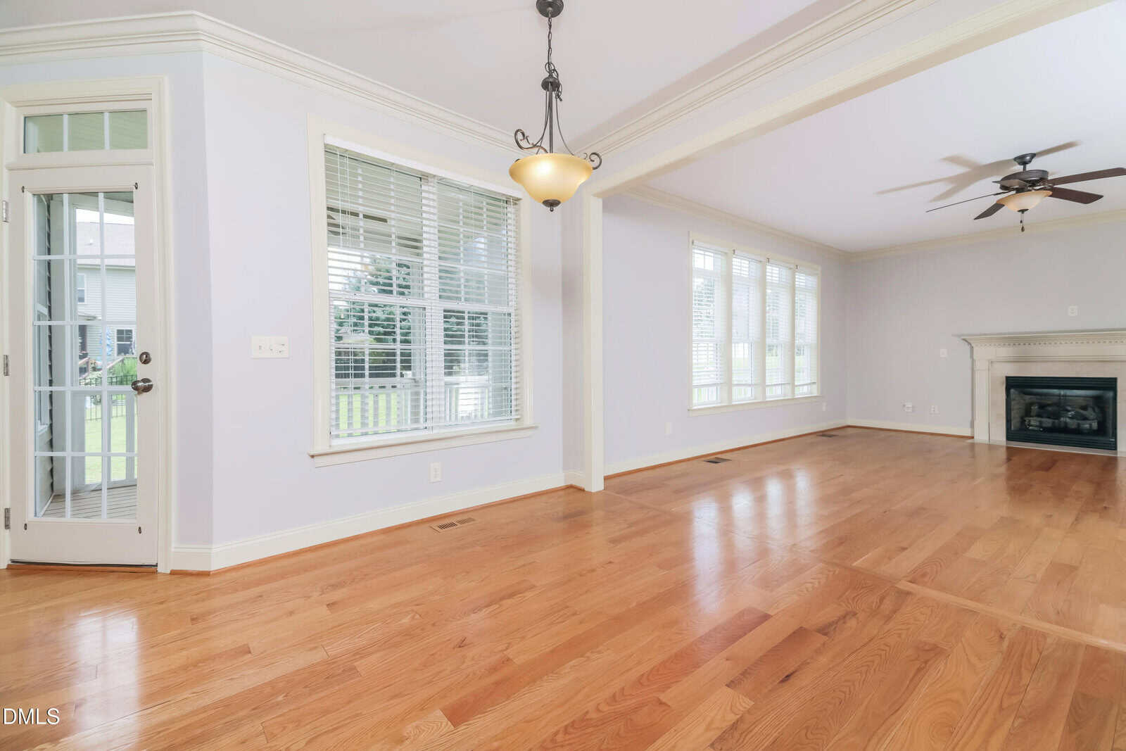Spacious, empty living room with light hardwood floors, large windows with white blinds, a ceiling fan, a hanging light fixture, a white fireplace, and a glass door leading outside. Walls are painted light gray.