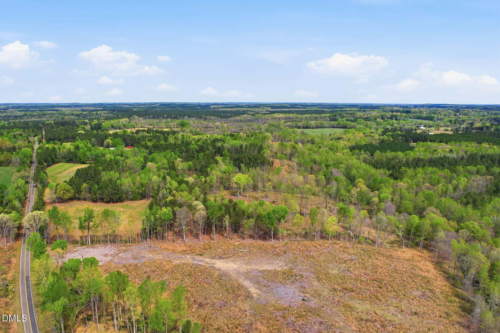 Aerial view of a rural landscape with a road on the left, open grassland, patches of trees, and forested areas under a blue sky with scattered clouds.