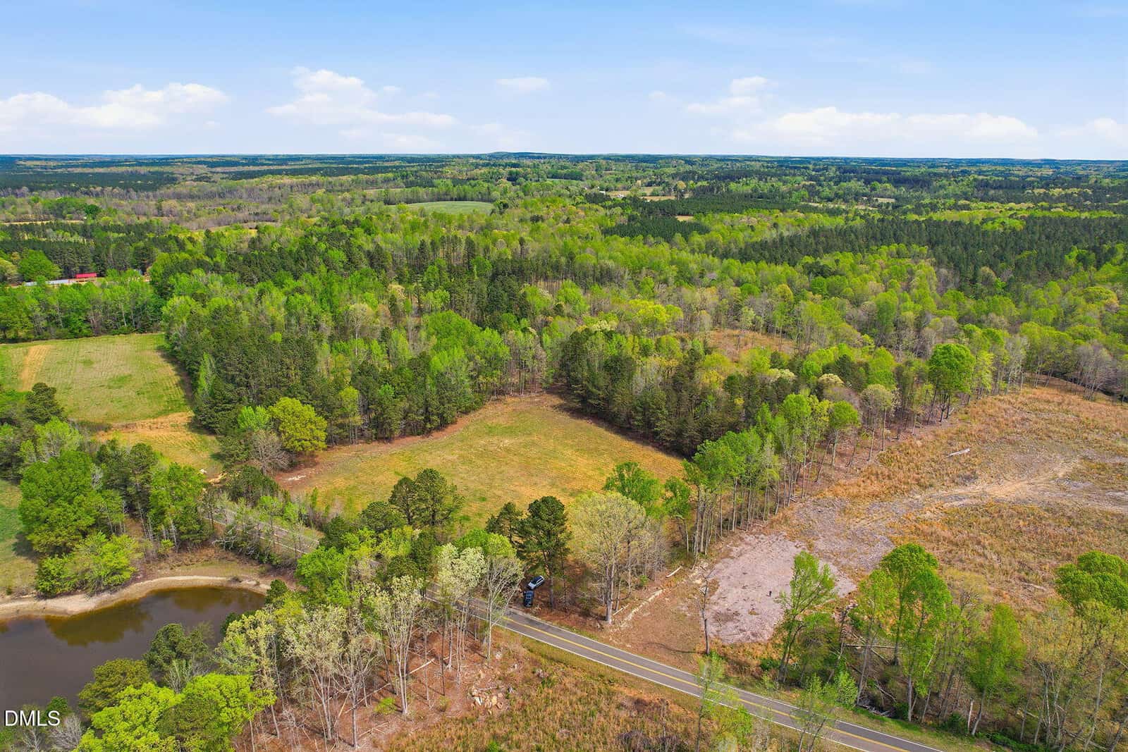 Aerial view of a rural landscape with a mix of forest, open fields, a pond in the foreground, and a paved road running through the scene under a blue sky with scattered clouds.