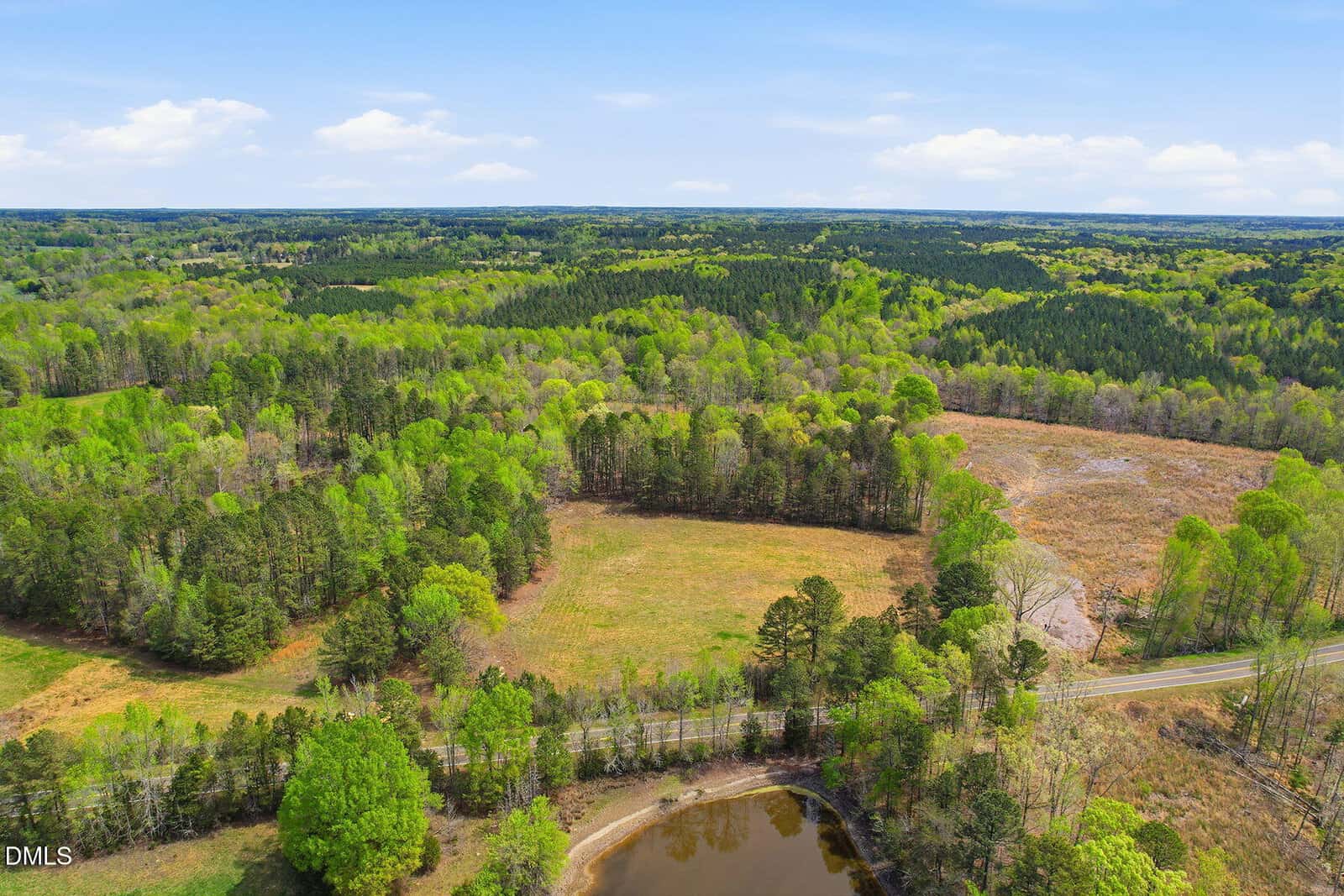 Aerial view of a rural landscape with a pond in the foreground, a road running alongside it, and wooded areas with green trees and open fields under a partly cloudy sky.