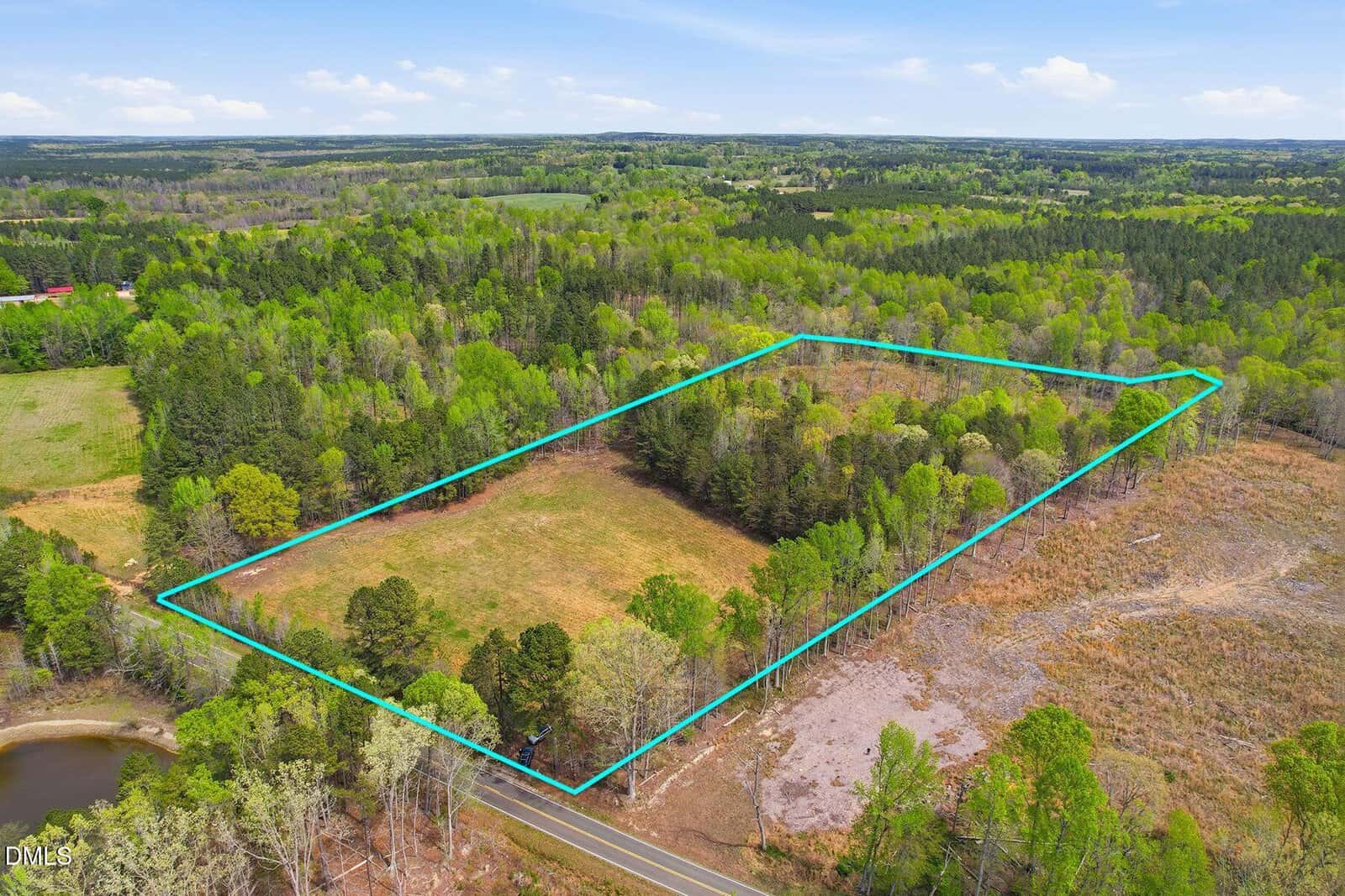 A rectangular plot of land outlined in blue sits next to a road, surrounded by trees, open fields, and a small pond in a rural area under a blue sky with scattered clouds.