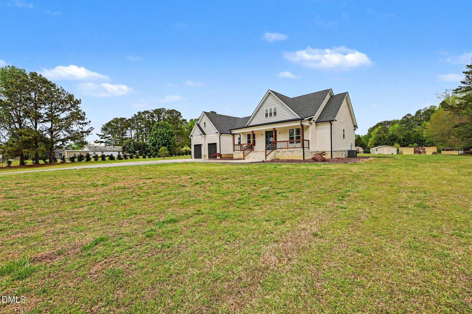 A single-story house with a covered front porch, three-car garage, and large grassy yard, surrounded by trees under a blue sky with scattered clouds.
