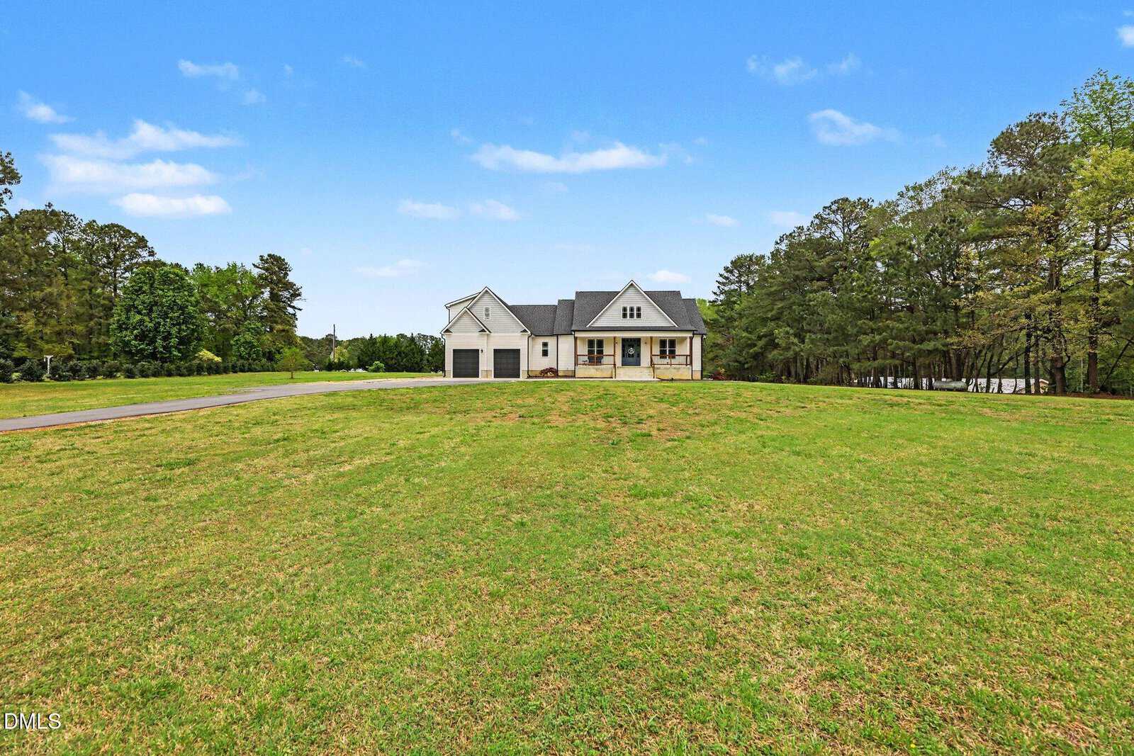 A single-story white house with dark roof sits on a large, open grassy lawn surrounded by trees under a bright blue sky with scattered clouds.