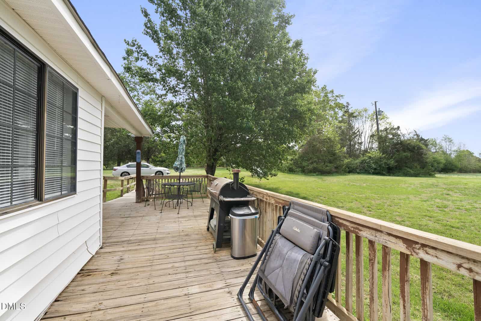 A wooden deck with a grill, chairs, table, and umbrella overlooks a grassy yard with trees. Part of a white house is visible on the left, and a white car is parked in the background.