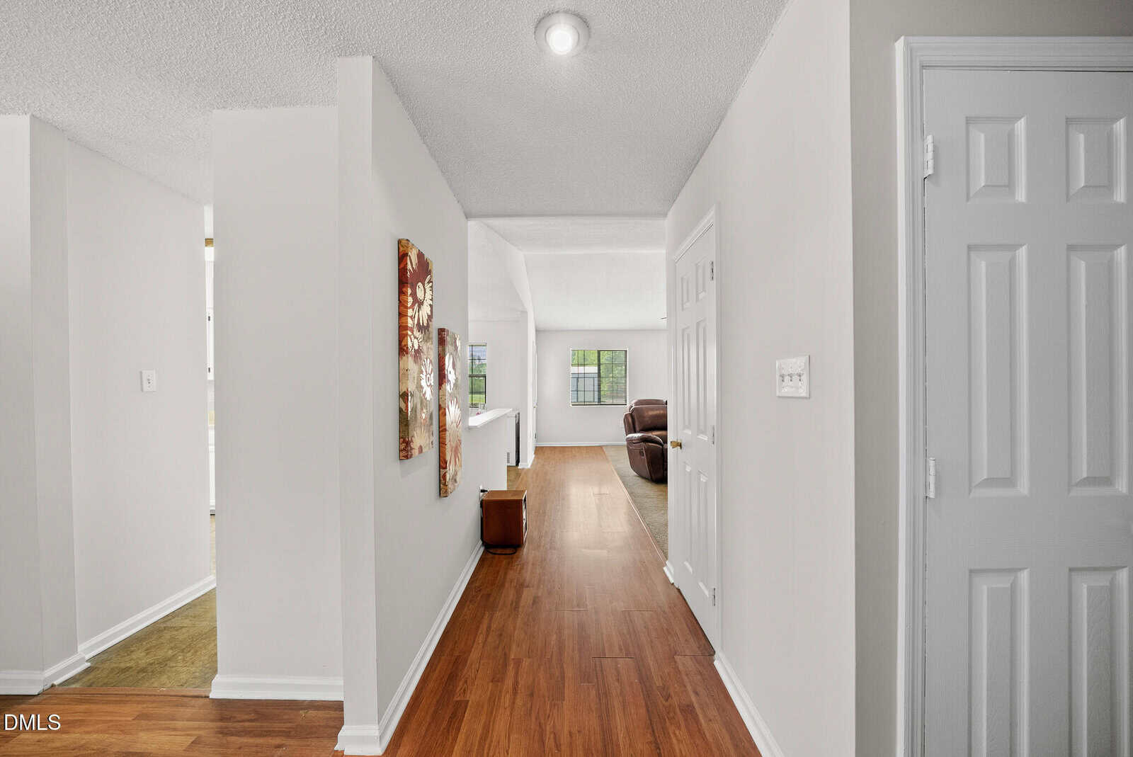A hallway with wood flooring leads to a bright living room. Two floral paintings hang on the left wall, and a small bench sits near the living area. White walls and doors create a clean, modern look.