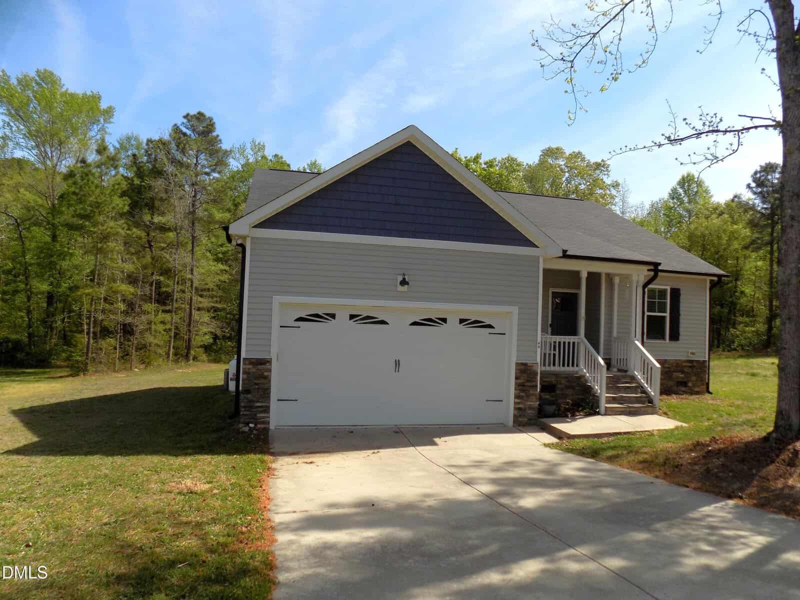 Charming single-story house at 60 Wickersham Way, Louisburg, featuring gray siding, a dark roof, attached two-car garage, white trim, and a small porch with steps. Surrounded by trees and greenery on a sunny day, this home offers both comfort and curb appeal.