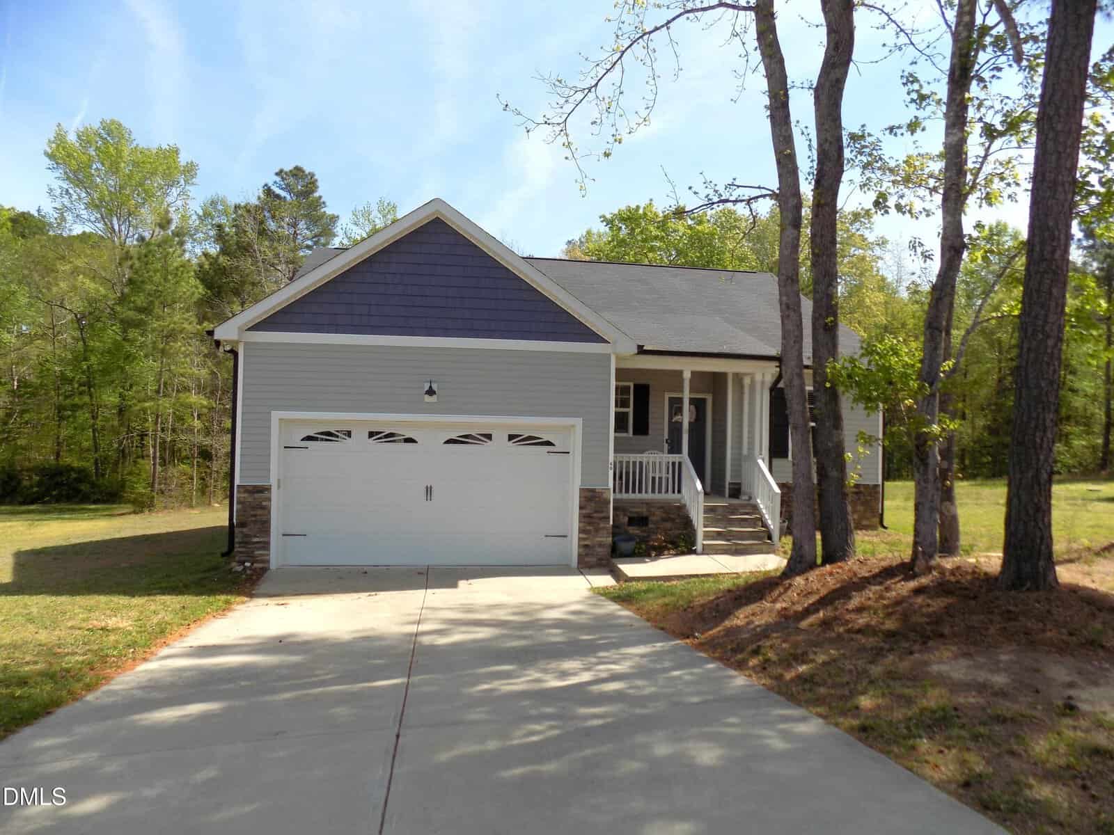 A single-story house with gray siding, a two-car garage, front porch, and steps. The home is surrounded by trees and has a wide concrete driveway leading to the garage.
