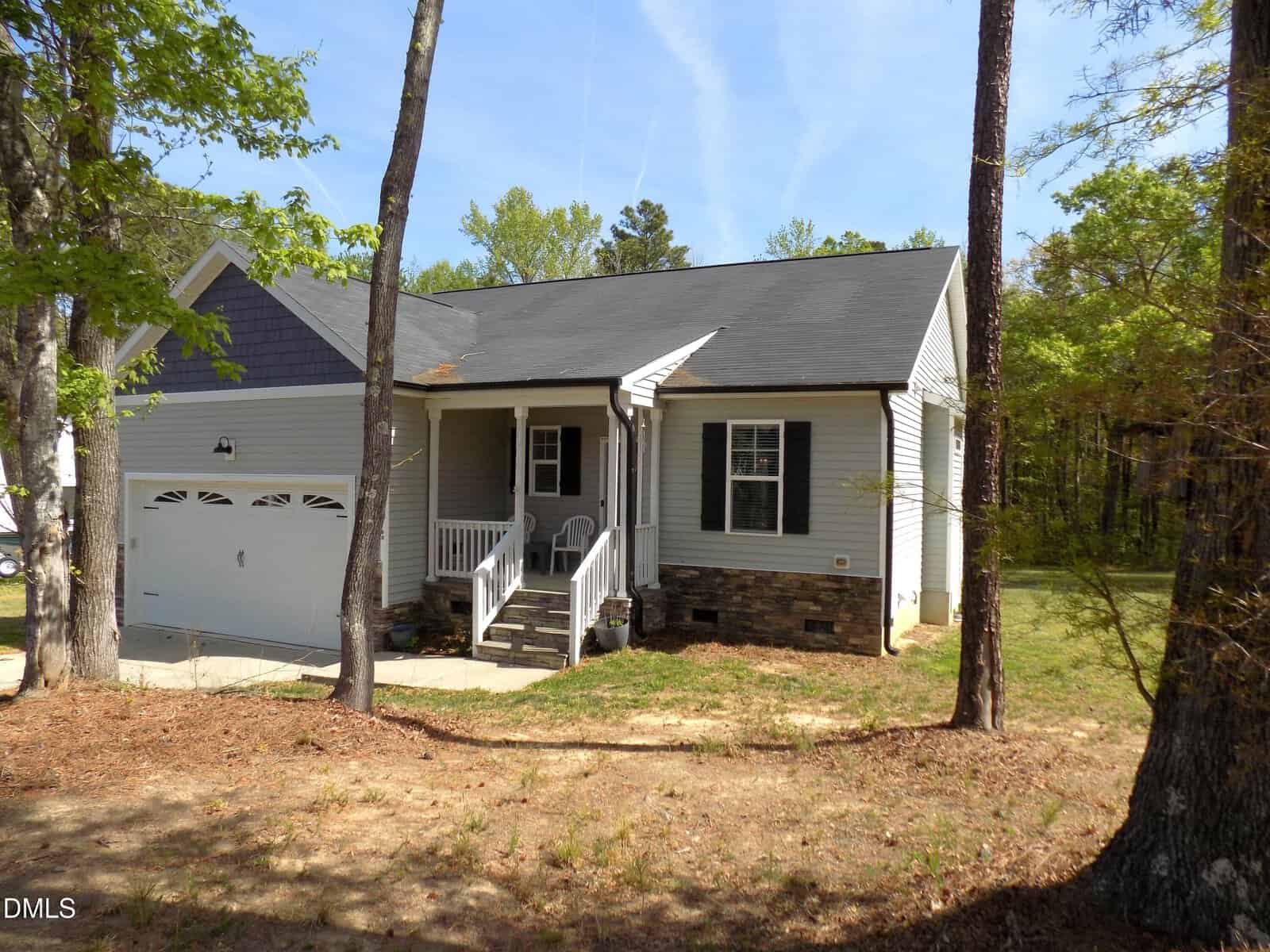 A small gray house with a front porch, white trim, and attached garage sits among tall trees on a sunny day. Steps lead up to the porch, and two chairs are visible near the front door.