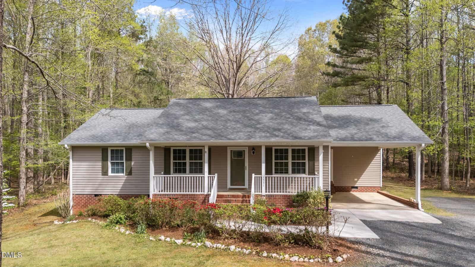 Single-story house with a gray roof, beige siding, white trim, and a welcoming front porch at 1551 Dogwood Drive in Oxford. The property features a carport on the right and a spacious driveway, surrounded by mature trees and a beautifully landscaped yard with shrubs and flowers.