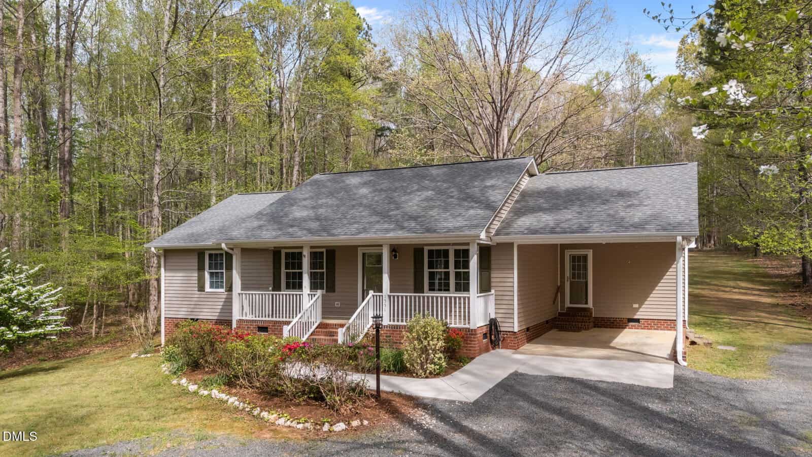 Single-story beige house with a covered front porch, white railings, brick foundation, and attached carport, surrounded by trees and a driveway leading up to the entrance.