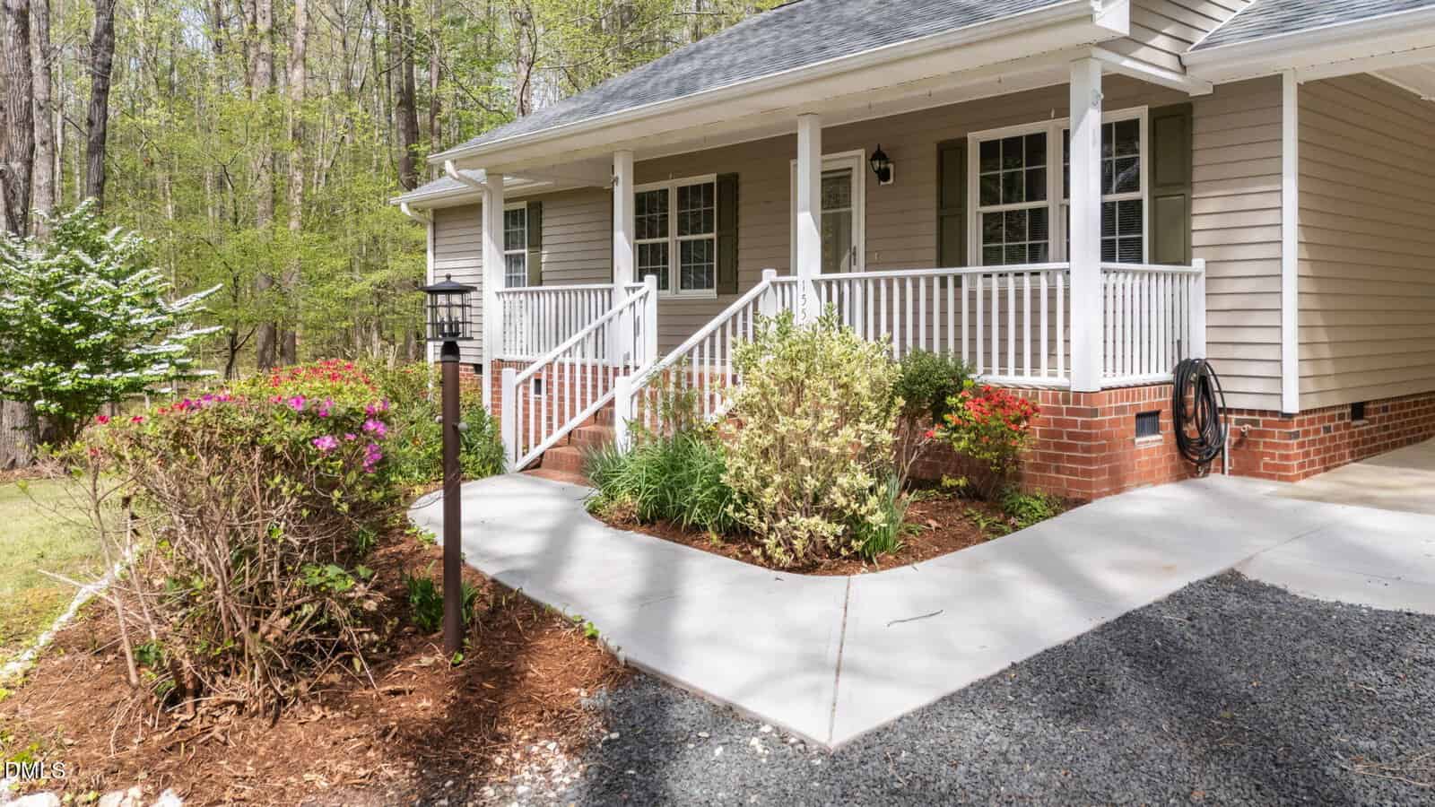 A single-story house with a covered porch, white railing, and brick foundation. There are shrubs, flowers, and a bird feeder along the concrete walkway leading to the front steps.