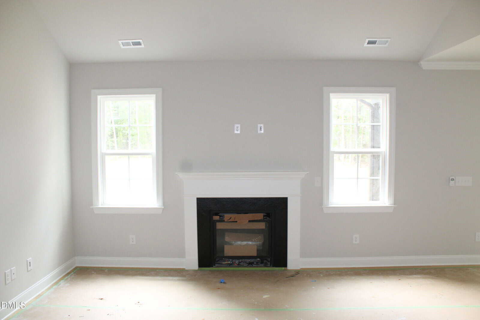 A bright, empty room with two tall windows and a white fireplace centered on a pale gray wall. The floor is unfinished, and the room appears to be under construction or renovation.