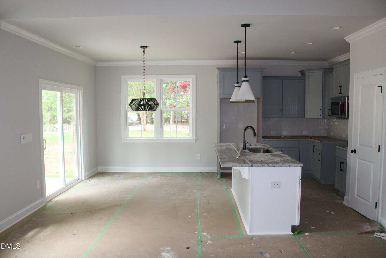 Unfurnished kitchen and dining area with gray cabinets, white island, pendant lights, and large windows. Floor is covered with protective material, and sliding glass doors open to a green backyard.