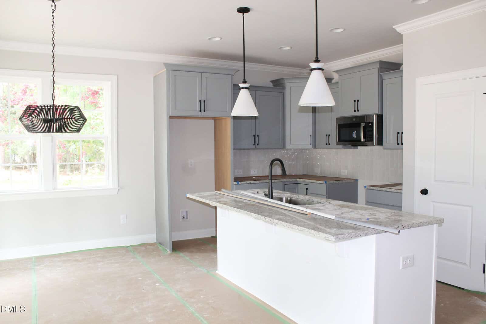 Modern kitchen with gray cabinets, white walls, two pendant lights over a large island, and a window letting in natural light. Floor is covered for protection, indicating ongoing construction or renovation.