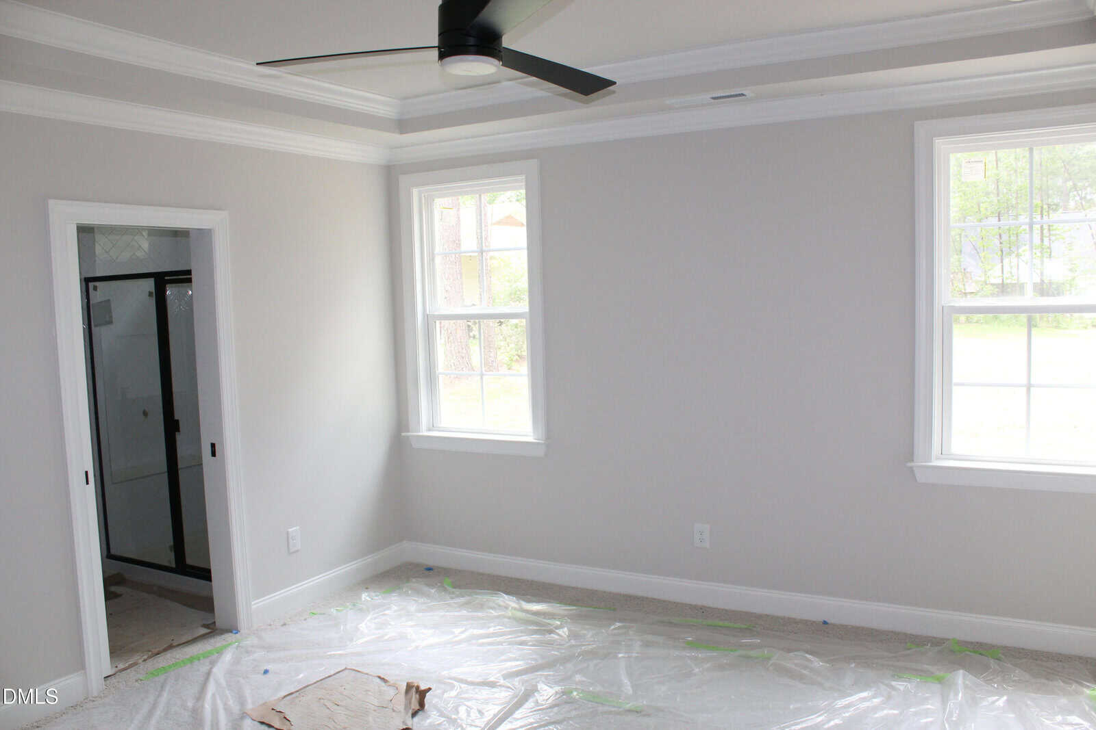 A bright, empty room with two windows, a ceiling fan, white walls, and an attached bathroom. The floor is covered with protective plastic sheets.