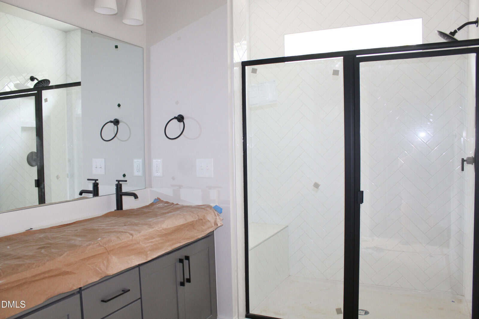 A modern bathroom with a glass shower enclosure, white tiled walls, a large mirror, and a vanity covered with brown protective paper. Black fixtures and a ring towel holder are visible.