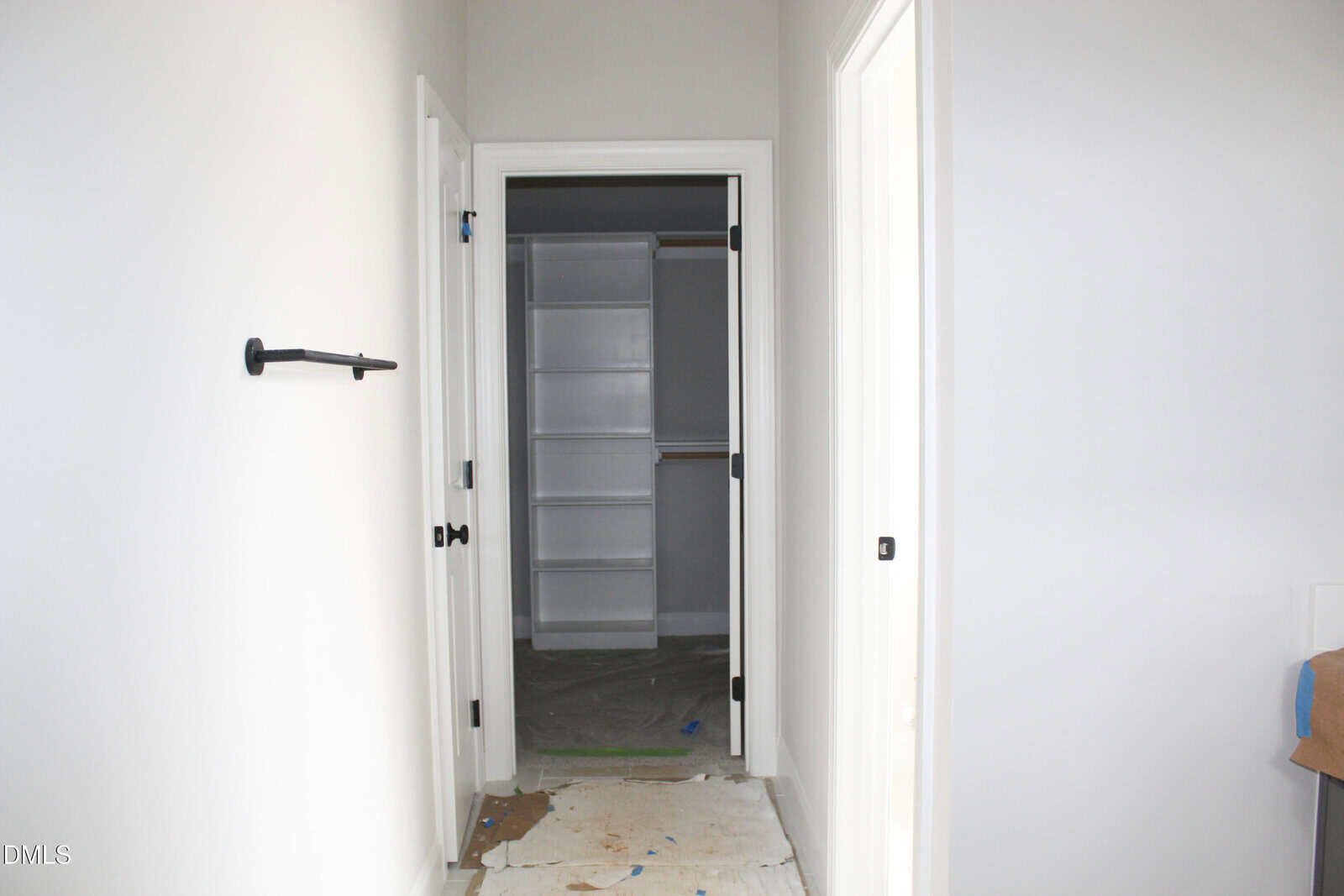 A hallway with light-colored walls leads to an open closet with white shelves. The floor is covered with protective paper, and doors are open on both sides. A towel bar is mounted on the left wall.