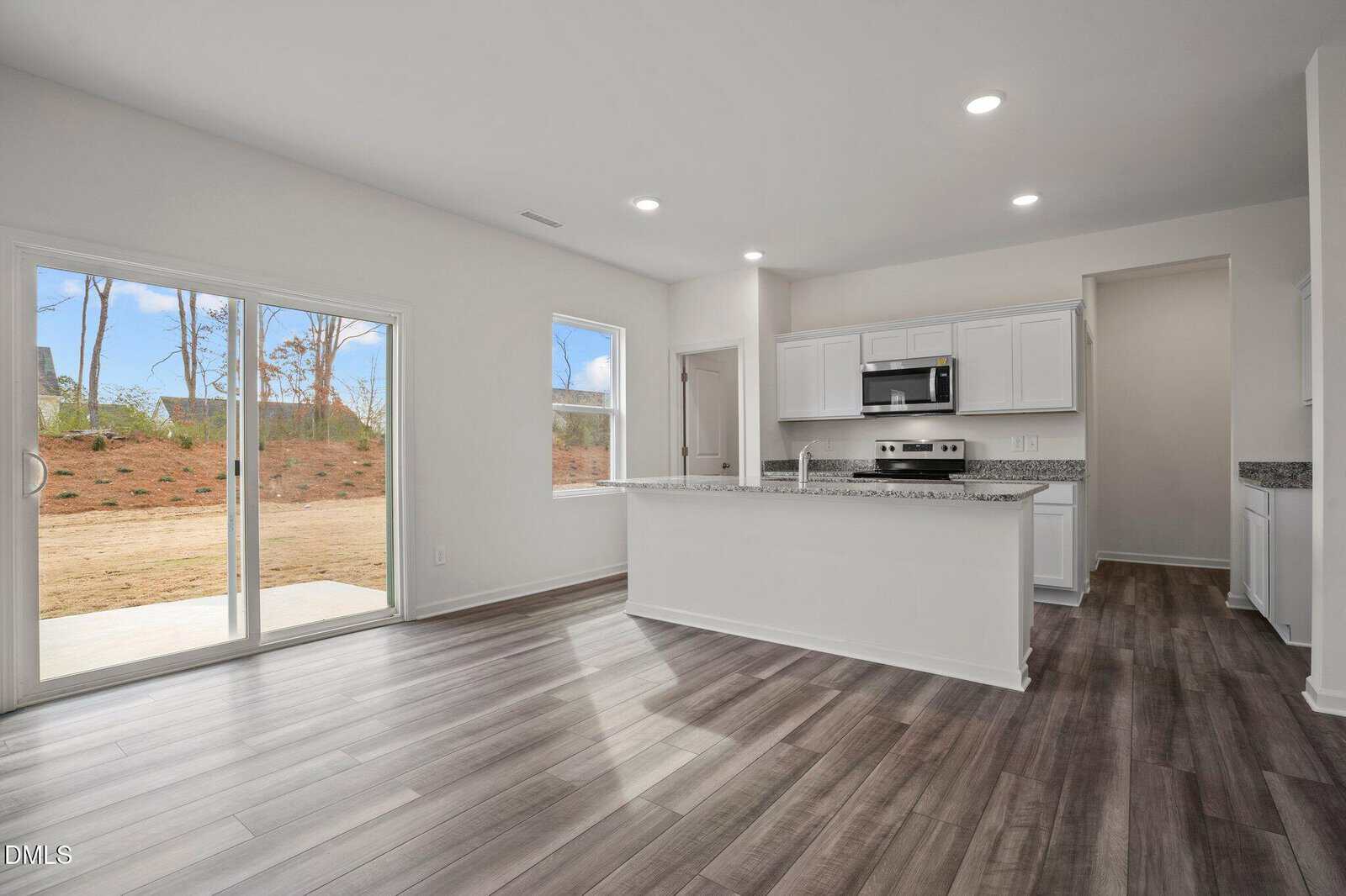 Modern kitchen with white cabinets, granite countertops, stainless steel appliances, and wood-look flooring. Large sliding glass doors and a window let in natural light, with a view of a backyard area.