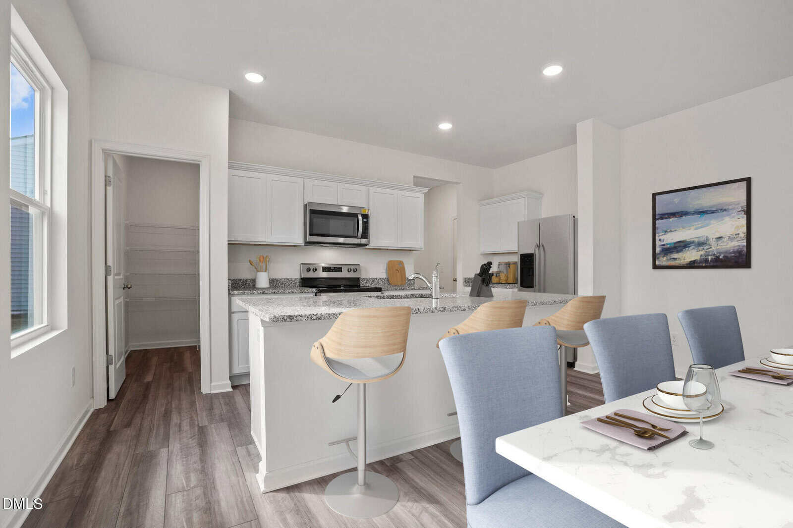 Modern kitchen with white cabinets, granite countertops, stainless appliances, and breakfast bar with three light wood stools. Dining table with blue chairs in foreground. Large window and artwork on the wall.
