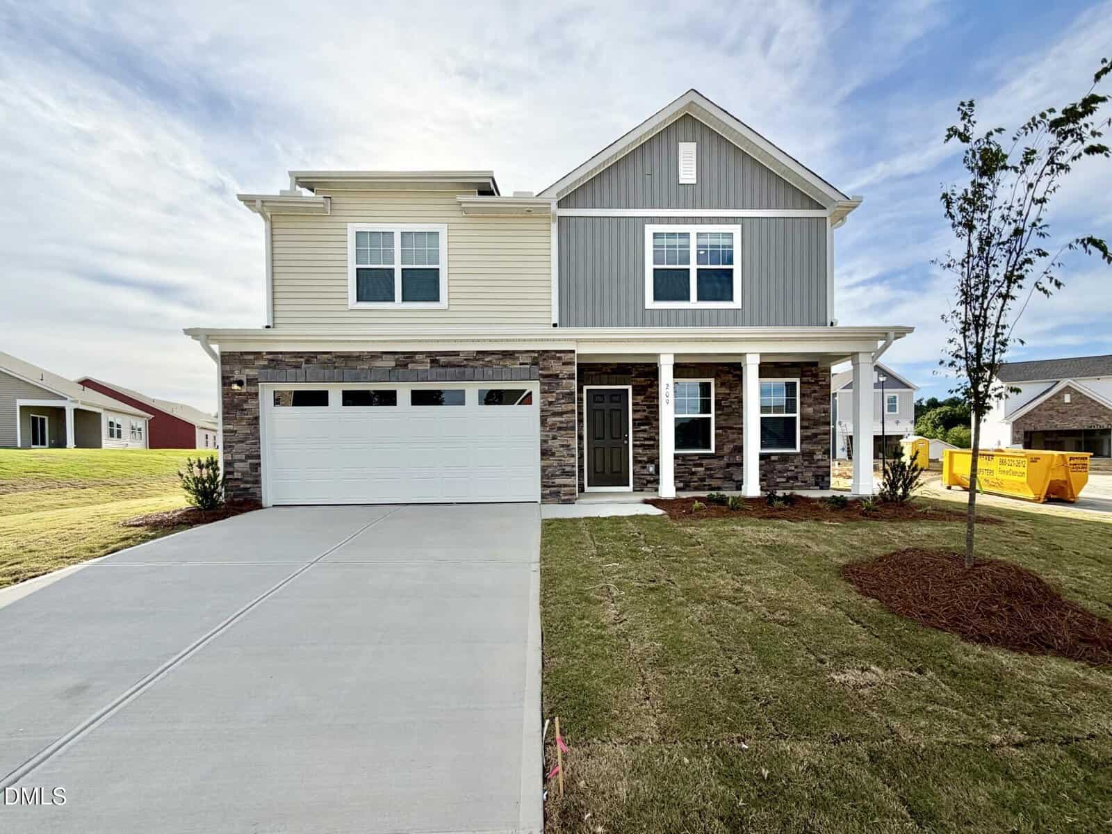 Two-story suburban house with beige and gray siding, a two-car garage, stone accents, a black front door, and a small front lawn. A yellow dumpster is visible on the right side near a tree.