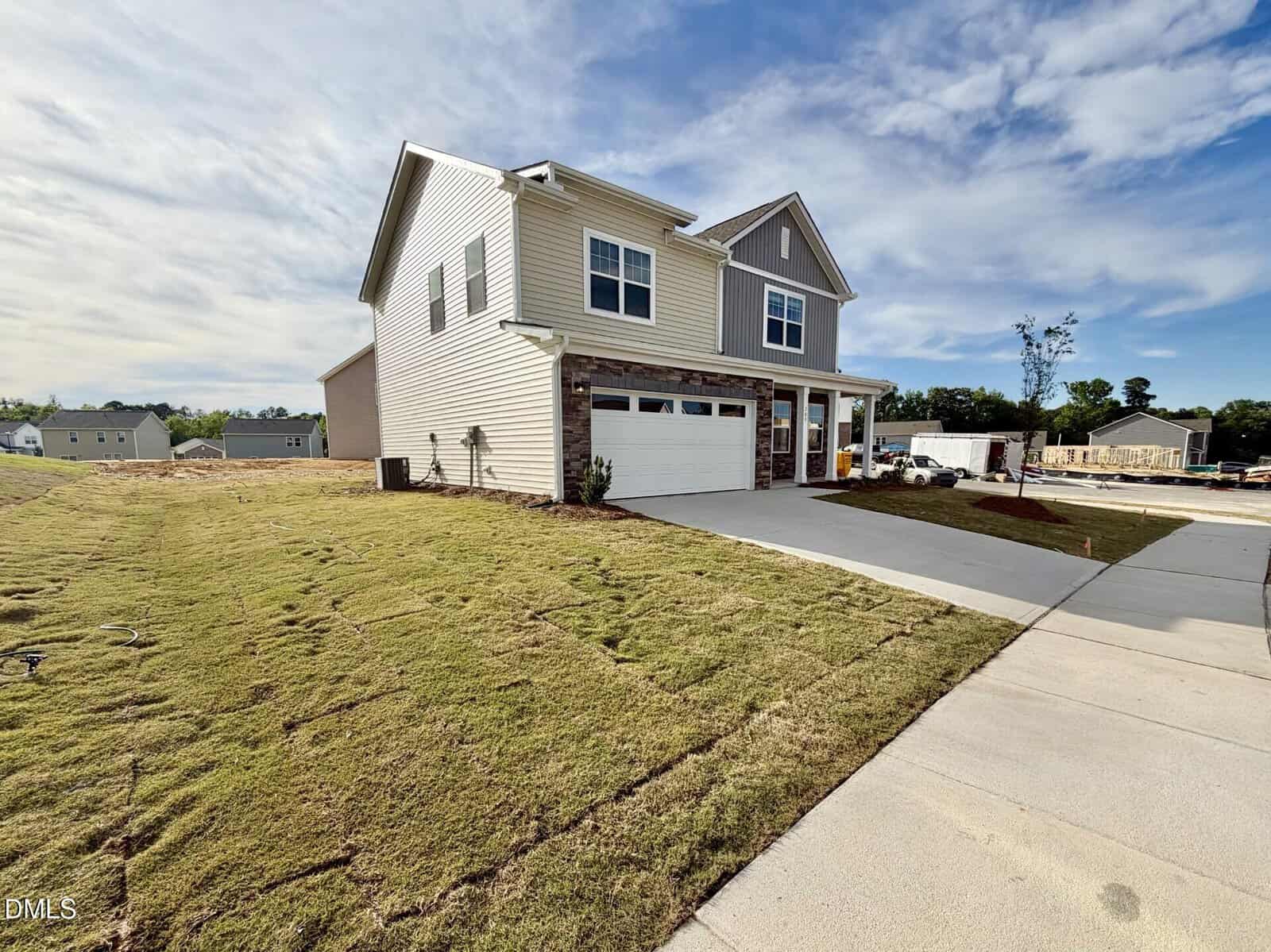 Two-story modern house with beige siding, stone accents, and a double garage. Newly laid sod covers the front yard, and a wide concrete driveway leads to the street. The sky is partly cloudy.