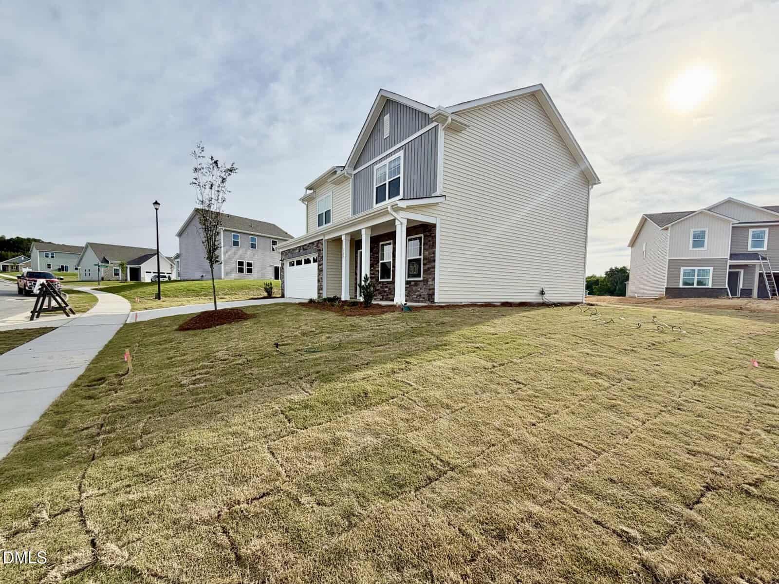 A newly constructed two-story house with beige siding, a front porch, and a two-car garage sits on a freshly sodded lawn in a suburban neighborhood under a partly cloudy sky.