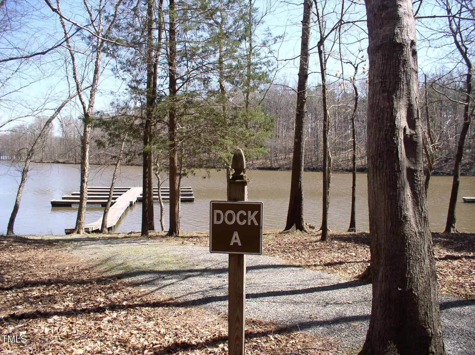 A brown sign labeled “Dock A” stands among leafless trees near a gravel path at 3003 Jonathans Landing, with a wooden dock extending into a calm, brownish lake in the background—an inviting scene in Bullock.