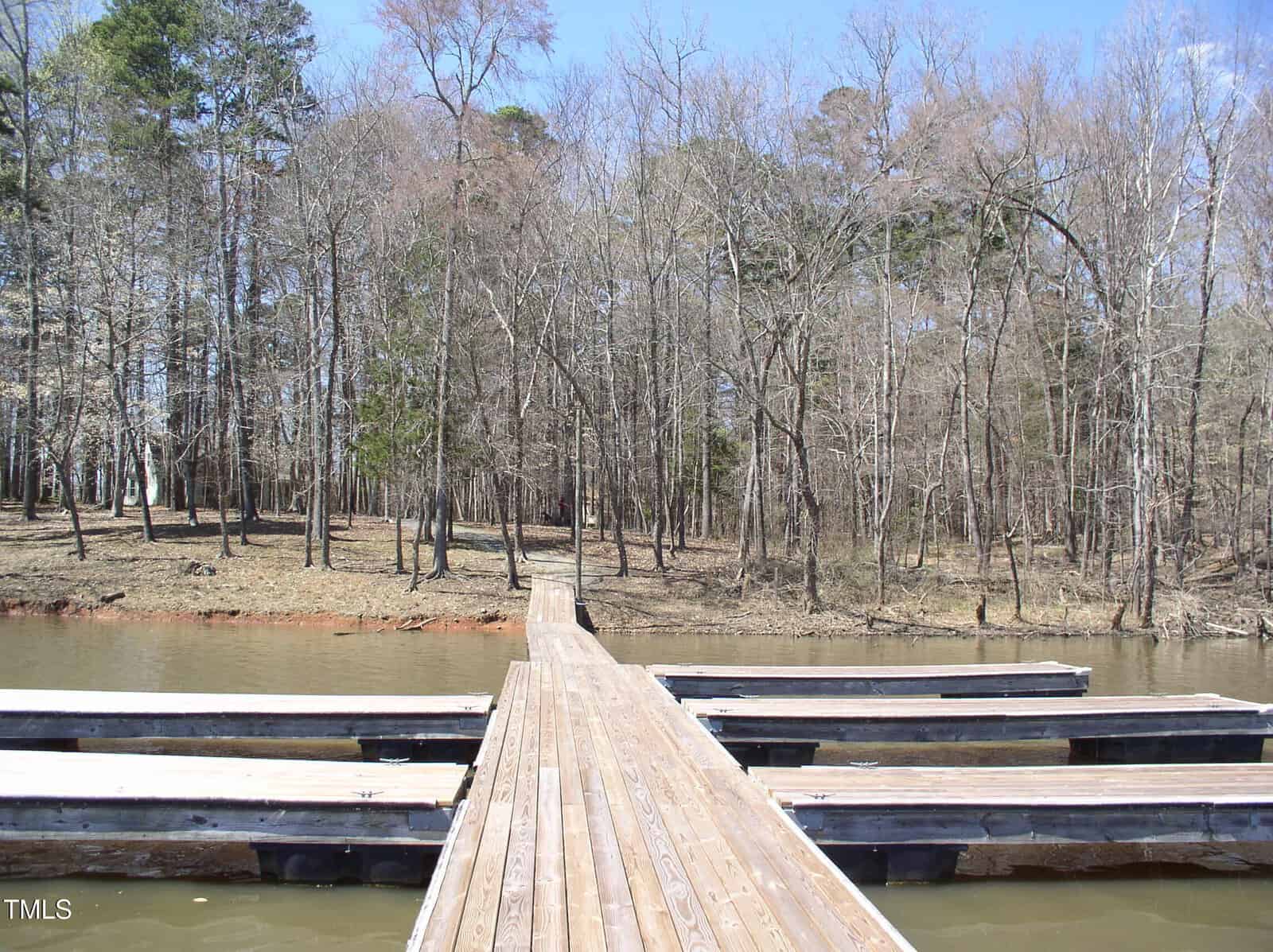A wooden dock extends over murky water toward a forest of leafless trees. Several empty boat slips are on either side of the dock. The sky is clear and blue.