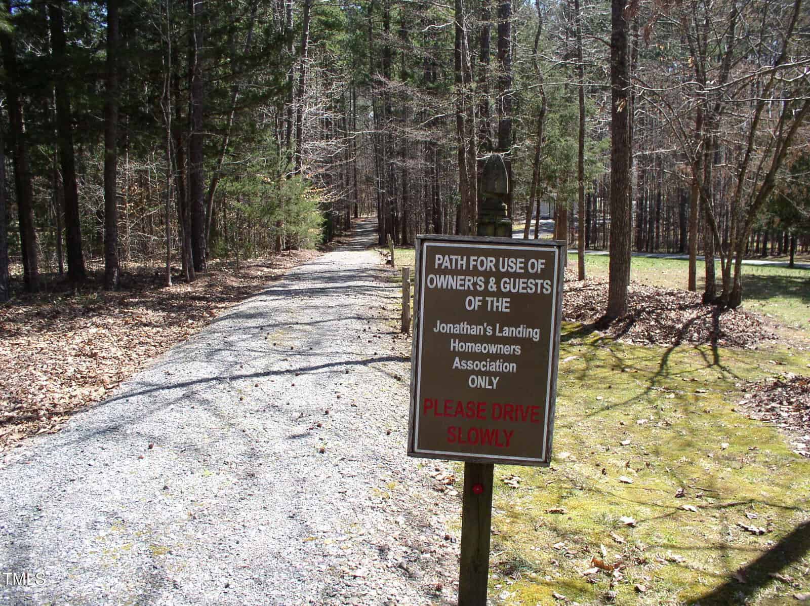 A gravel driveway bordered by trees, with a sign stating the path is for Jonathan’s Landing Homeowners Association owners and guests only. The sign also instructs drivers to proceed slowly.