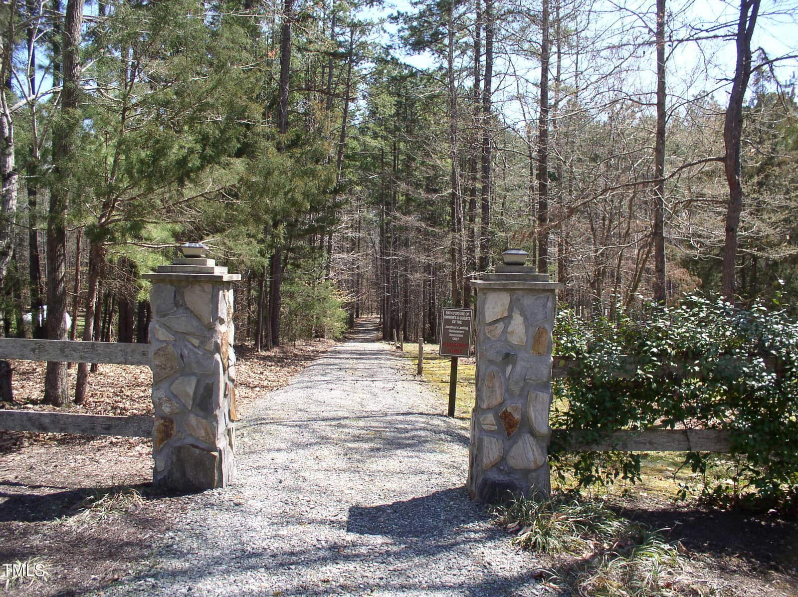 A gravel path passes between two stone pillars and wooden fences, leading into a wooded area with tall trees. A brown sign is on the right pillar, and sunlight filters through the branches.
