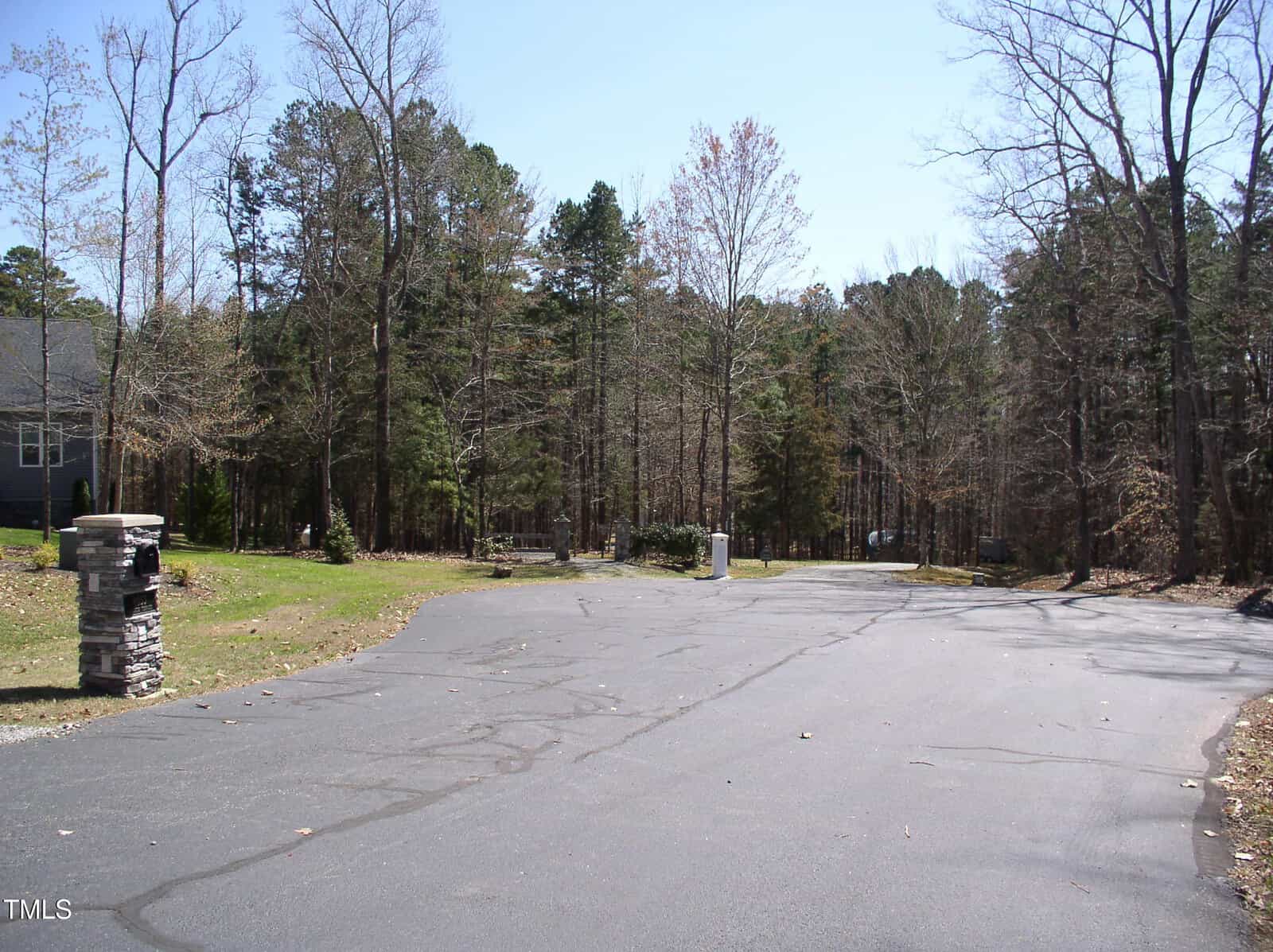A paved road curves through a wooded suburban area with leafless trees and some evergreens. A stone mailbox and a house are partially visible on the left side of the image. The sky is clear and blue.