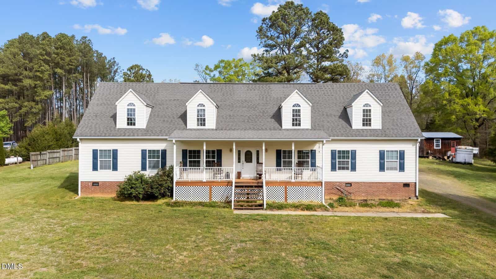 Large white house with gray roof, multiple dormer windows, front porch with white railings, brick foundation, and grassy yard surrounded by trees under a blue sky with scattered clouds.