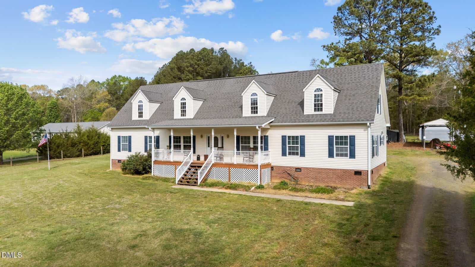 A large white two-story house with dormer windows, a covered front porch, and brick foundation sits on a spacious lawn with surrounding trees under a blue sky with scattered clouds.