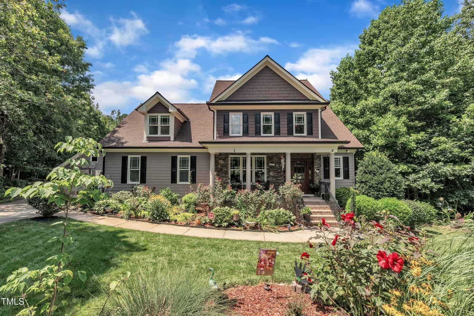 Two-story suburban house with brown siding and a covered front porch, surrounded by a well-maintained garden with a variety of flowers, bushes, and trees under a partly cloudy sky.